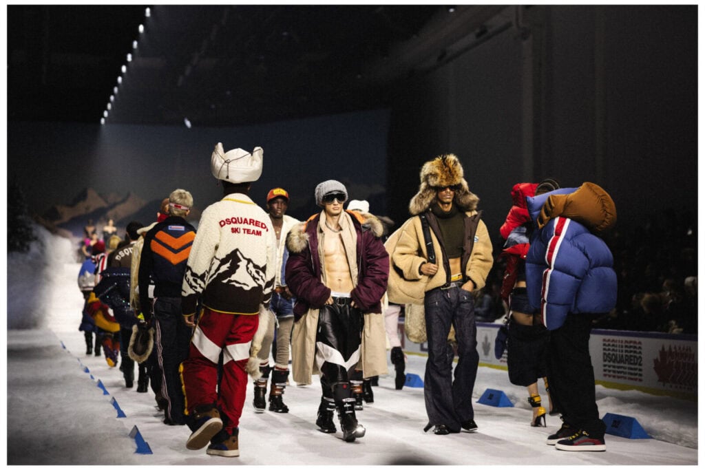 Models walk down a runway wearing colorful, winter-themed outfits and ski gear. The scene resembles an indoor ski slope, with bright lights, snowy decor, and spectators watching the fashion show.
