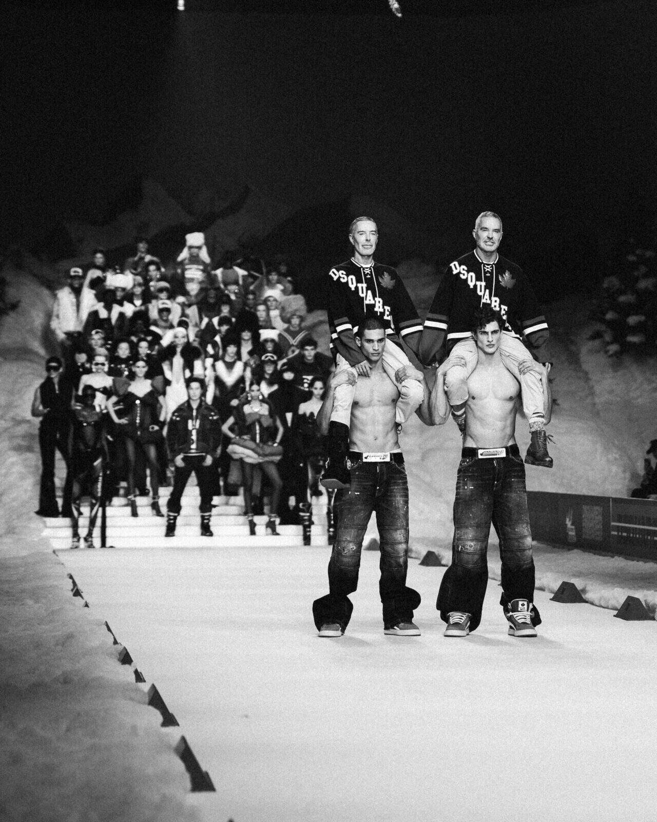 Black and white photo of the Dsquared2 AW26 runway show: two shirtless male models carry others on their shoulders, all in jeans and “Dsquared” jerseys. A large group poses dramatically on the steps in the background.