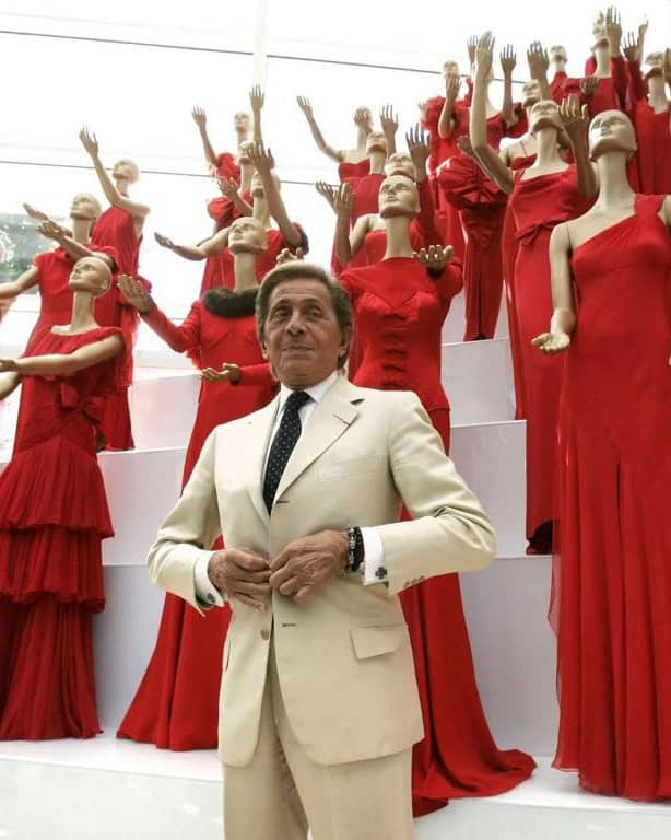 Valentino Garavani in a cream suit stands in front of mannequins dressed in elegant red gowns, arranged in rows on white steps, with their arms raised dramatically. The background is brightly lit by natural light.