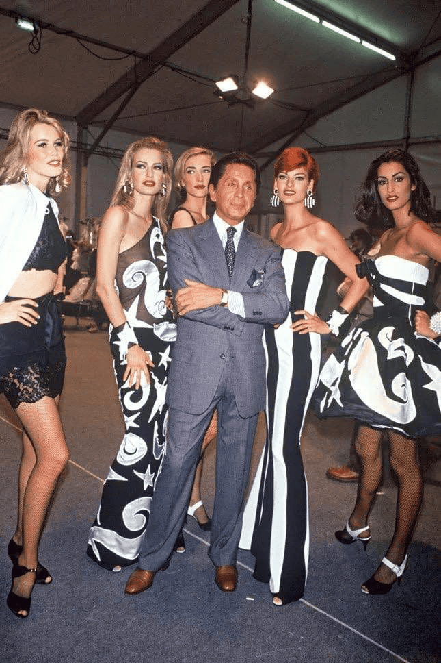 Valentino Garvani in a suit stands confidently with arms crossed, surrounded by five women in bold black and white patterned dresses and dramatic earrings, posing together under a tent with bright lights.