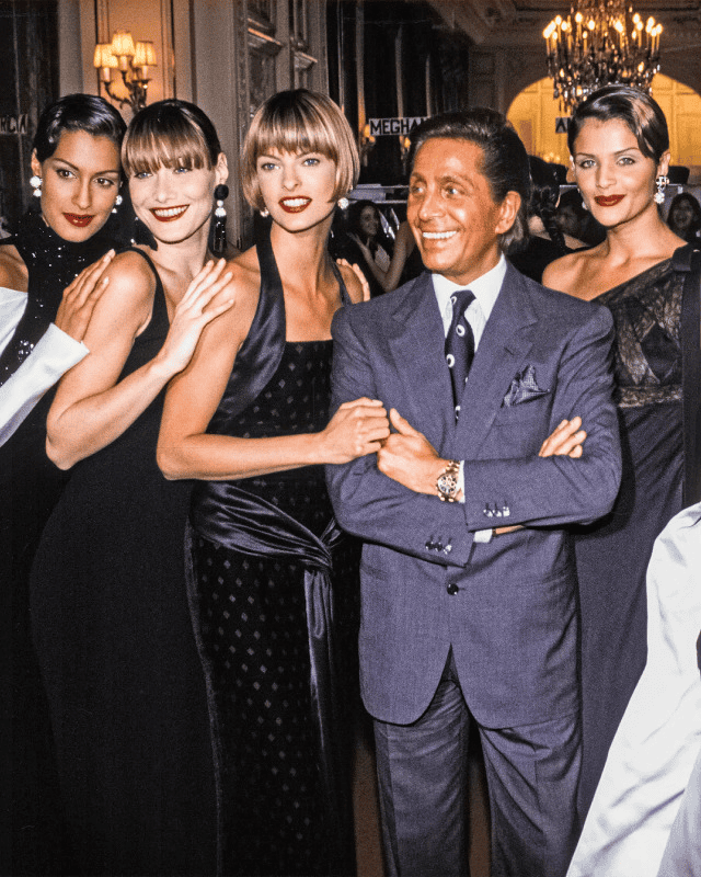 A group of four women in elegant black dresses pose with a smiling Valentino Garavani in a suit at a glamorous indoor event with chandeliers and a crowd in the background.