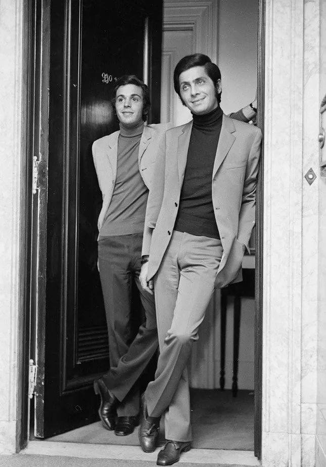 Valentino Garavani and a friend in suits and turtlenecks stand relaxed in a doorway. One man leans against the doorframe with arms crossed; the other stands slightly in front with legs crossed, smiling. The setting appears to be indoors.