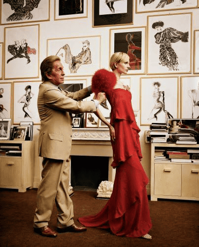 Valentino Garavani in a tan suit adjusts the back of a womans flowing red dress in a room with framed fashion sketches covering the wall. The woman stands poised in front of a fireplace, surrounded by books and cabinets.