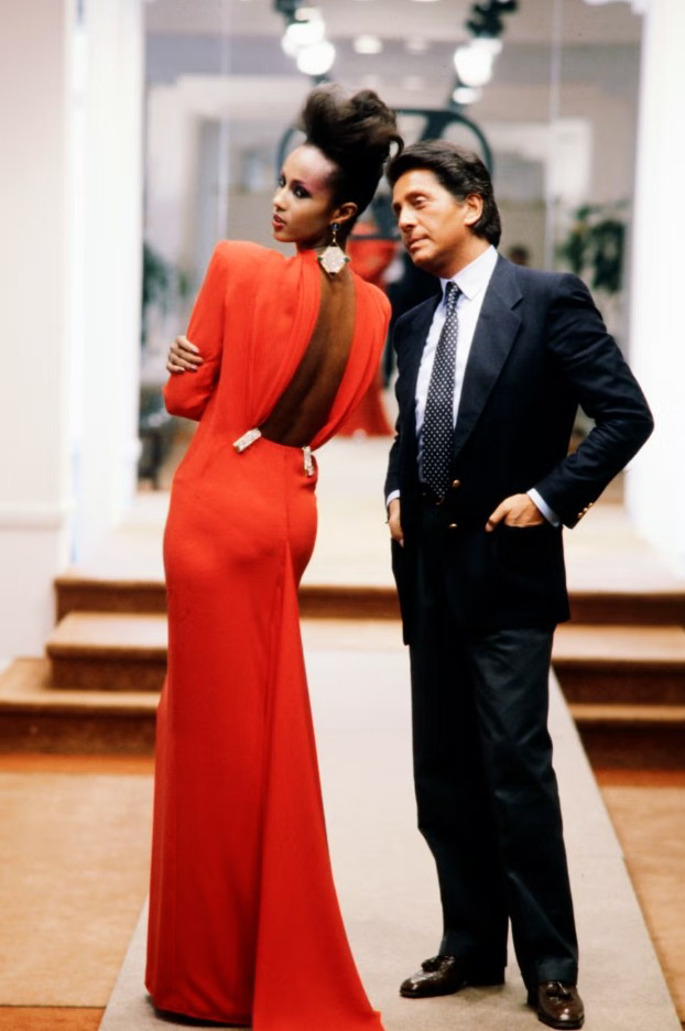 A woman in a long, backless red dress poses confidently next to Valentino Garavani in a dark suit and tie, standing in a stylish indoor setting with a runway and mirrors in the background.