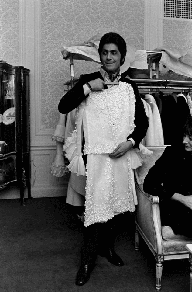 Valentino Garavani in formal attire stands in a room holding a white, embellished dress on a hanger, smiling. Behind him is a clothing rack with similar garments and ornate wallpaper. Another person is seated nearby.