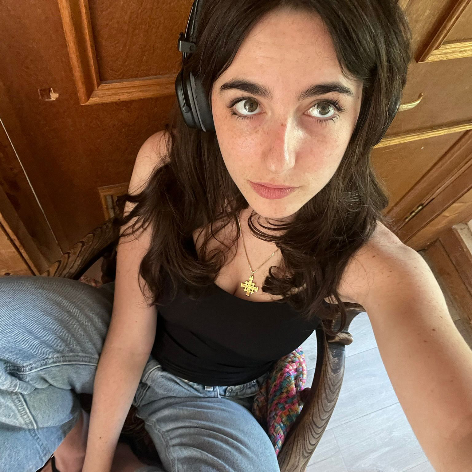 A woman with long brown hair, part of a cold team, wears headphones, a black tank top, and jeans while sitting on a chair indoors. She looks up at the camera from above, with a wooden door visible in the background.