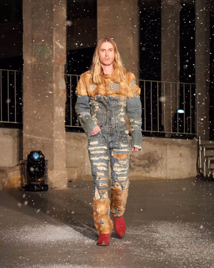 A model with long hair walks on a snowy AW26 runway wearing a tan outfit with a fur vest, matching pants, and lace-up boots, set against an industrial concrete backdrop.