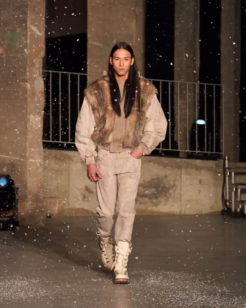 A model with long brown hair walks the AW26 runway in a fur vest layered over a beige jacket and pants, paired with tall white boots. Snow appears to fall in the industrial, concrete-walled setting.
