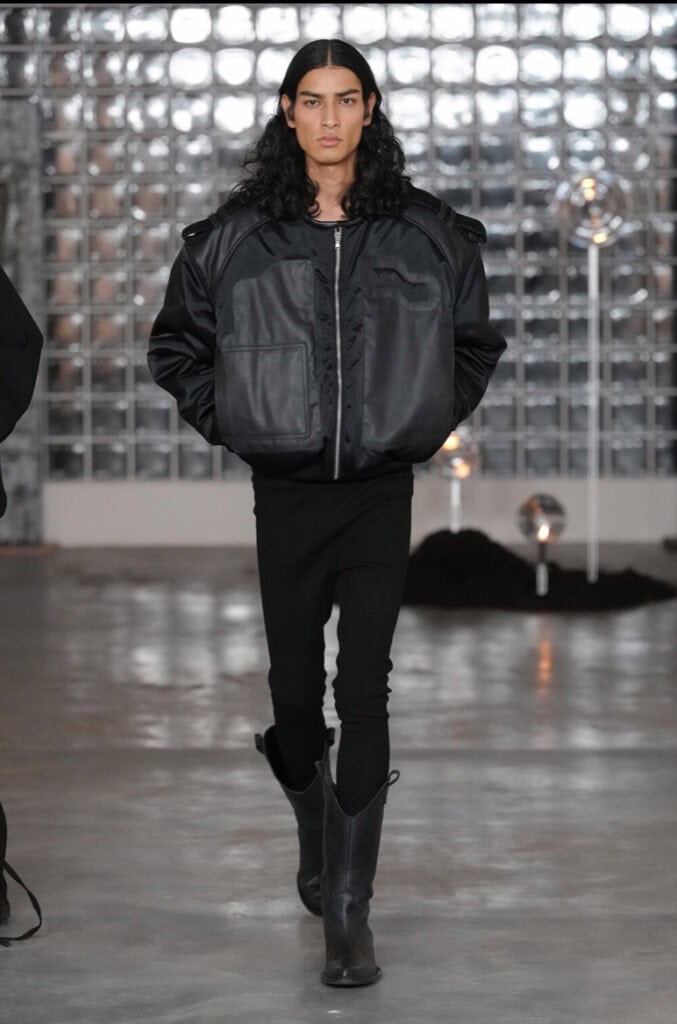 A model with long dark hair walks on the AW26 runway, wearing a black oversized leather jacket, black pants, and black cowboy boots. The background features glass brick walls and dim industrial lighting.