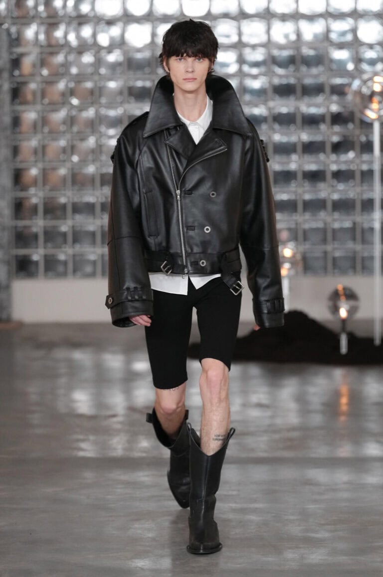 A model walks the AW26 runway in an oversized black leather jacket, white shirt, black knee-length shorts, and black boots. The backdrop features a wall of glass blocks and a polished concrete floor.