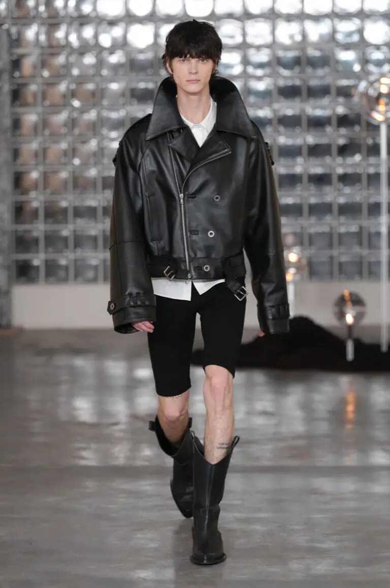 A model walks the AW26 runway in an oversized black leather jacket, white shirt, black knee-length shorts, and black boots. The backdrop features a wall of glass blocks and a polished concrete floor.