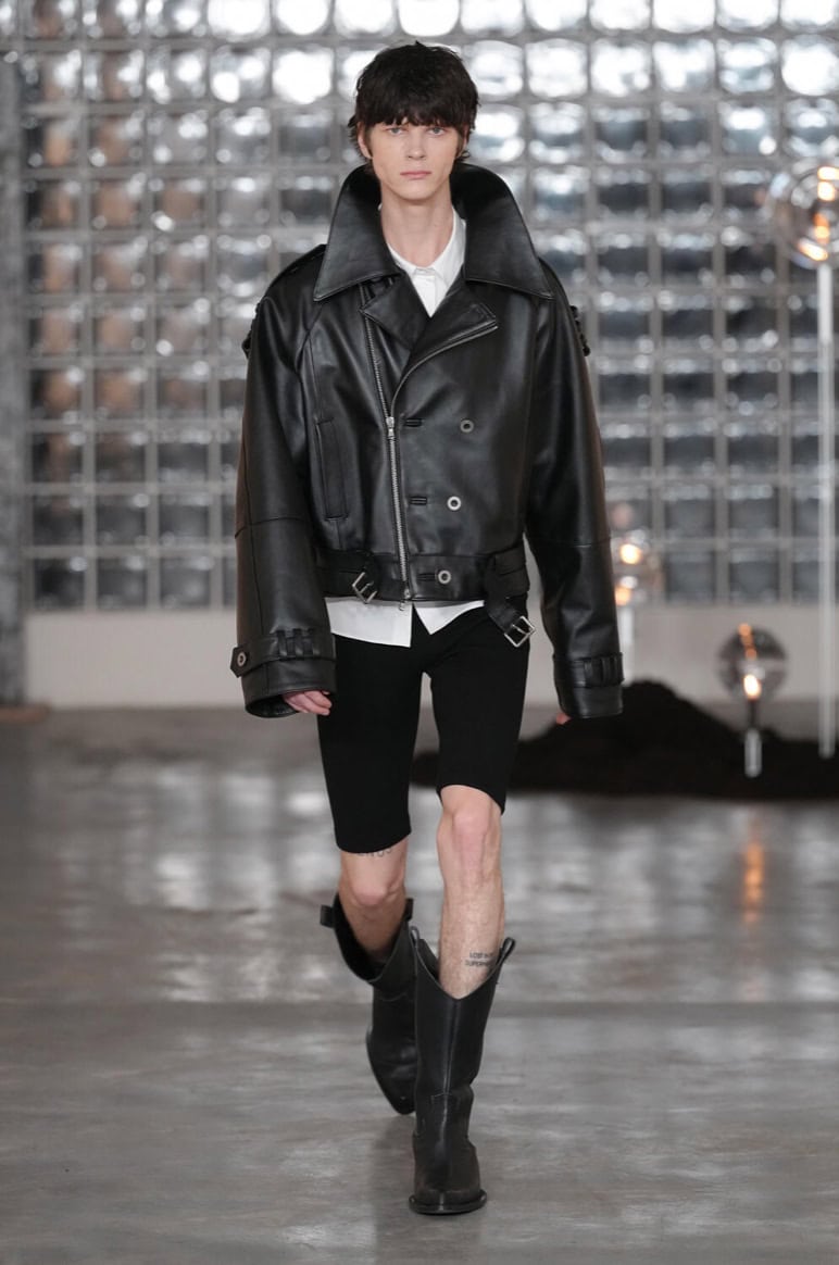 A model walks the AW26 runway in an oversized black leather jacket, white shirt, black knee-length shorts, and black boots. The backdrop features a wall of glass blocks and a polished concrete floor.