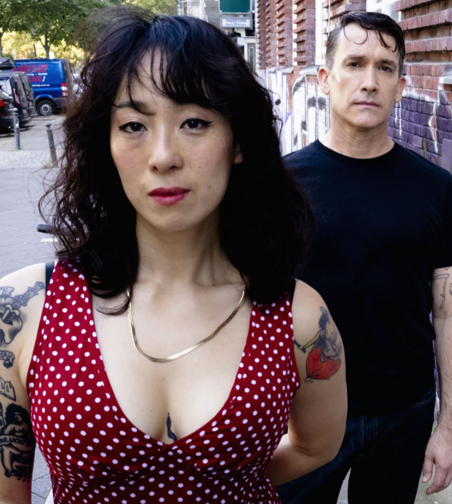 A woman with dark hair in a red polka dot dress stands in the foreground, looking at the camera, while a man in a black t-shirt stands behind her on a city sidewalk with brick walls and graffiti.