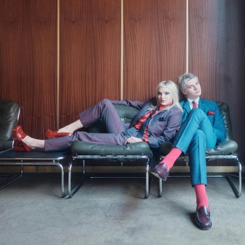 Two people in colorful suits sit on green chairs against a wood-paneled wall; one reclines in a purple suit, red shirt, and loafers, while the other sits upright in a blue suit with red socks, both with serious expressions.