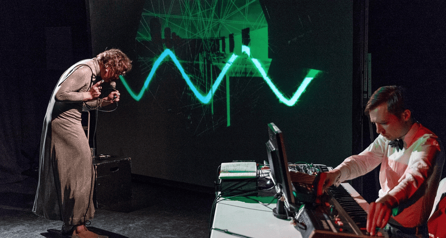 Harry Josephine Giles sings into a microphone on stage while another operates a keyboard and electronic equipment. Green wave-like graphics are projected onto a screen behind them.