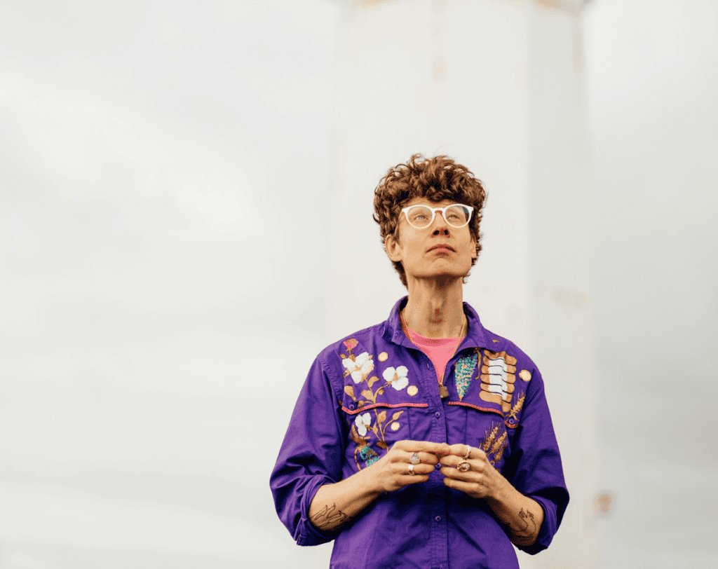 Michael Pedersen: The current Edinburgh Makar (Poet Laureate), Michael weaves between English and Scots, crafting irreverent sketches of the modern Scottish cultural scene. Revisit his debut collection,Play With Me, or his most recent: The Cat Prince & Other Poems. stands outdoors, wearing a bright purple shirt with colorful embroidered designs, looking slightly upward with a neutral expression. The background is blurred and light-colored.