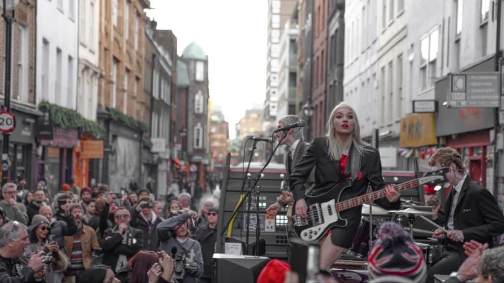 A band performs live on a busy city street, surrounded by a large crowd. The female bassist in front wears a black outfit and red tie, while many people watch and take photos, and tall buildings line the street.