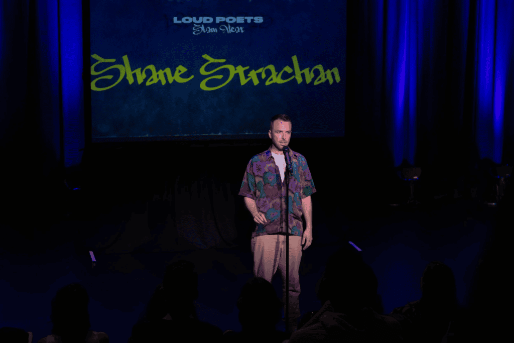 Shane Strachan stands on stage with a microphone, performing under blue lighting. Behind him, a screen displays the words LOUD POETS Slam West and Shane Strachan in bold yellow-green text.