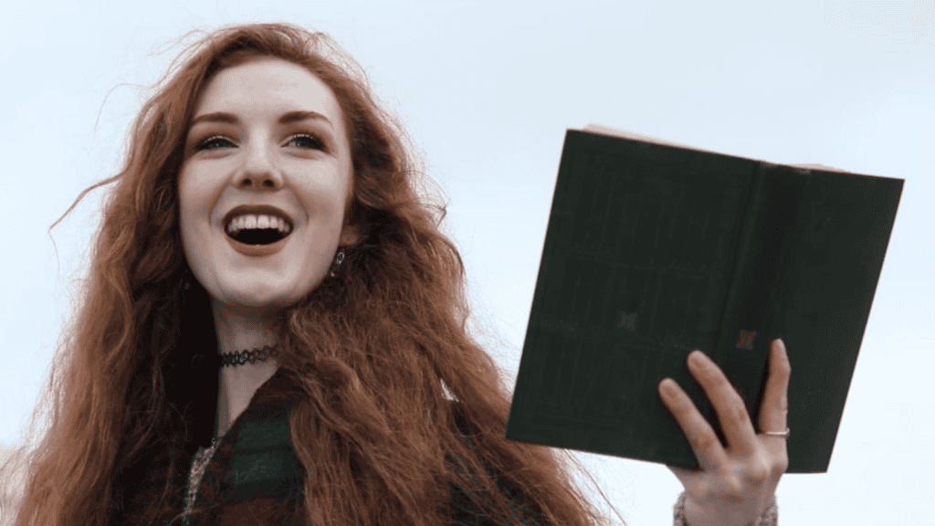 Len Pennie with long red hair smiles broadly while holding up an open green book outdoors. She wears a plaid scarf and a black choker necklace against a cloudy sky background.