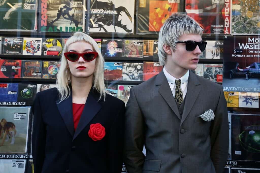 Two people with blonde hair and sunglasses stand in front of a wall covered with vinyl record covers. Both wear suits; the person on the left has a red rose pinned to their jacket, and the person on the right wears a tie and pocket square.