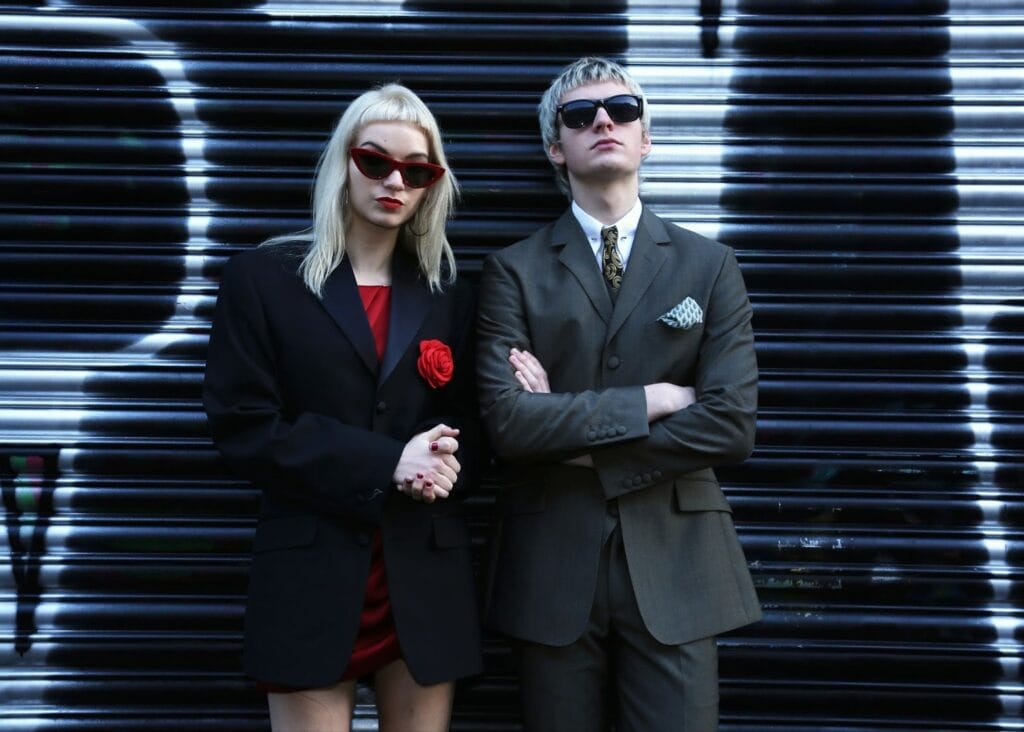 Two people channel The Molotovs: the Teenage Siblings Rocking Britain, wearing dark sunglasses and suits as they pose confidently in front of a black-and-white graffiti-covered shutter, blending rock energy with sharp style.