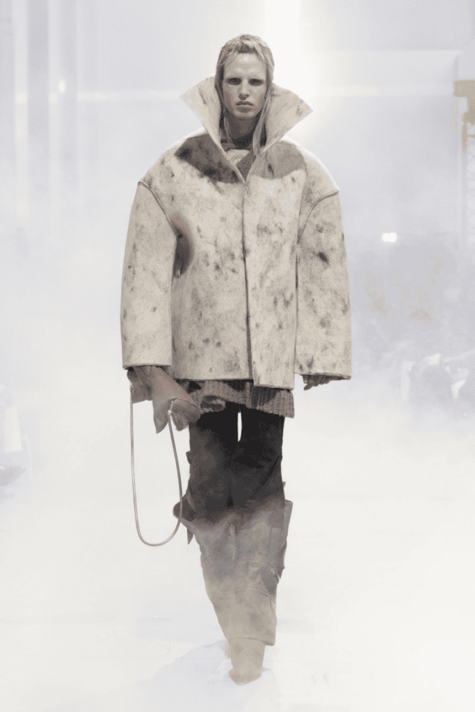A model walks through mist wearing an oversized, high-collared, off-white jacket with dark spots, layered over textured clothing, black pants, and tall slouchy boots on a runway.
