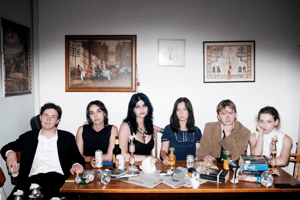 Six people sit at a cluttered table with drinks, newspapers, books, and figurines. They face forward with serious expressions. Framed art hangs on the wall behind them, and the lighting is dramatic, casting shadows on the white wall.