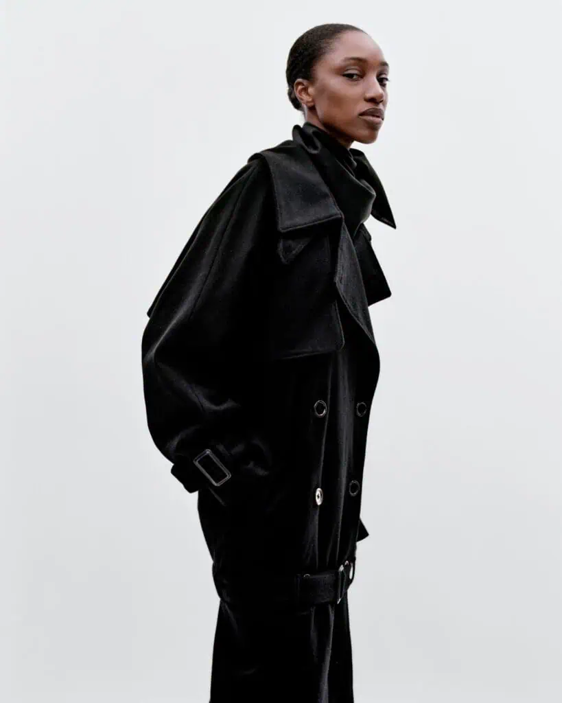 A person stands against a plain light background, wearing a long, black double-breasted trench coat with wide lapels and a high collar. Their expression is calm, and they are facing slightly to the left.