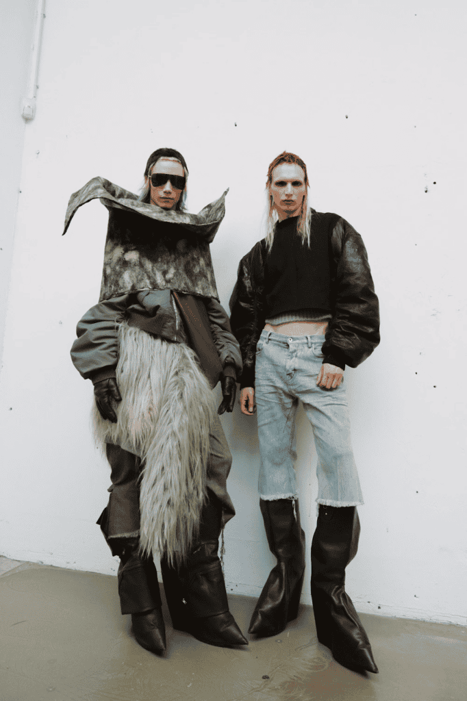 Two models stand against a white wall. One wears a large, sculptural, fur and fabric outfit with sunglasses and gloves. The other wears a black cropped jacket, light blue jeans, and oversized black boots.