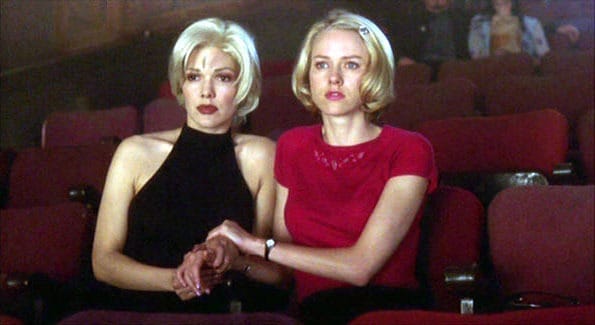 Two women with short blonde hair sit closely together in red theater seats, holding hands and looking ahead with serious expressions. The theater is mostly empty, with a few people visible in the background.