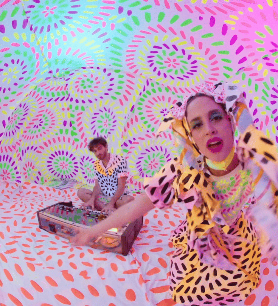 Two people in colorful, patterned outfits are inside a matching vibrant, patterned room. One person sits at a table with electronics; the other is closer to the camera, gesturing energetically toward the lens.