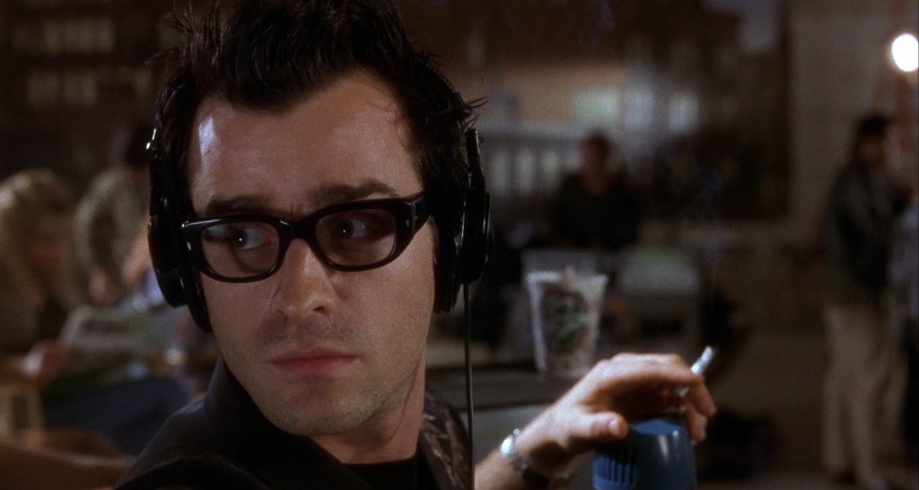 A man with dark hair and glasses wears headphones and looks to his left while holding a cigarette and a blue object. The background is a dimly lit, busy cafe with people sitting and talking.