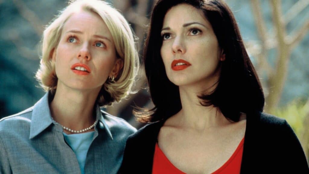 Two women stand close together outdoors, looking upward with serious, concerned expressions. One has short blonde hair and wears a light blue shirt and pearl necklace; the other has medium-length dark hair and wears a red top with a black sweater.