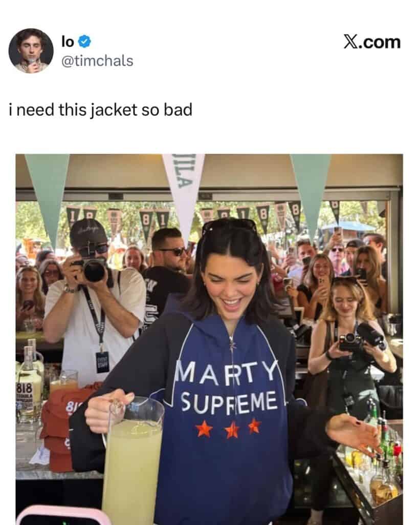 A woman smiles while pouring a drink behind a bar, wearing a blue jacket that says MARTY SUPREME with stars. A crowd of people behind her take photos and watch.