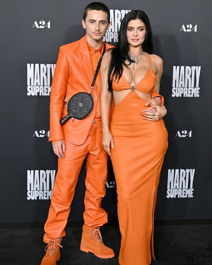 A man and woman pose on a red carpet, both dressed in matching bright orange outfits. The man wears a leather suit and boots, while the woman wears a strapless gown. The backdrop features the text Marty Supreme and A24.