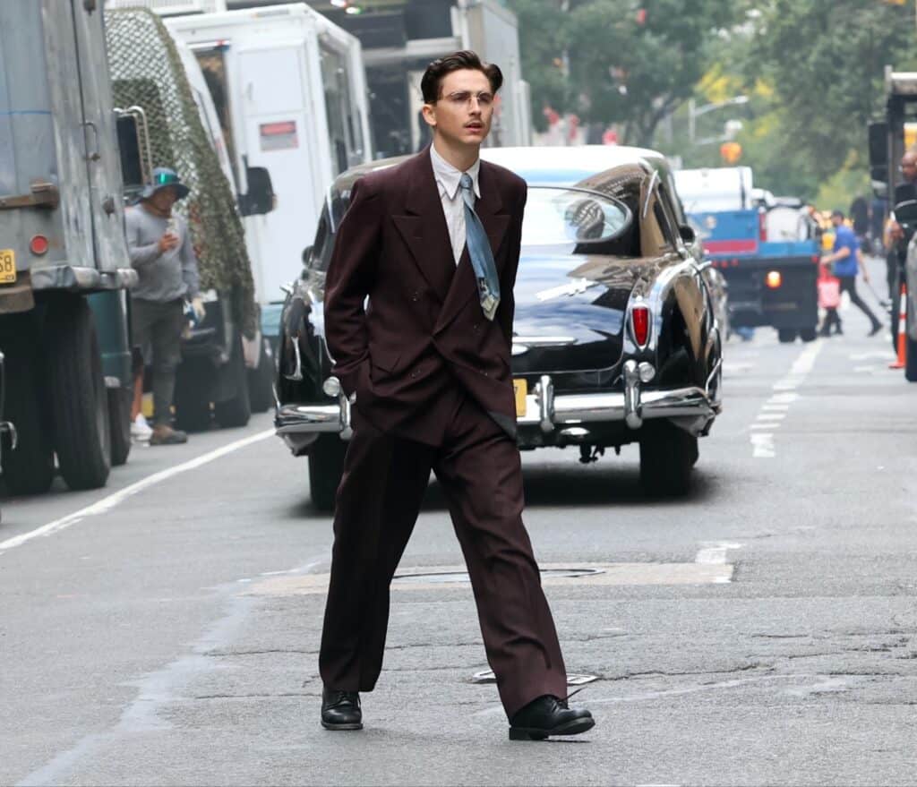 A man in a vintage dark brown suit and tie walks confidently across a city street, with classic cars and trucks parked along the road and people visible in the background.