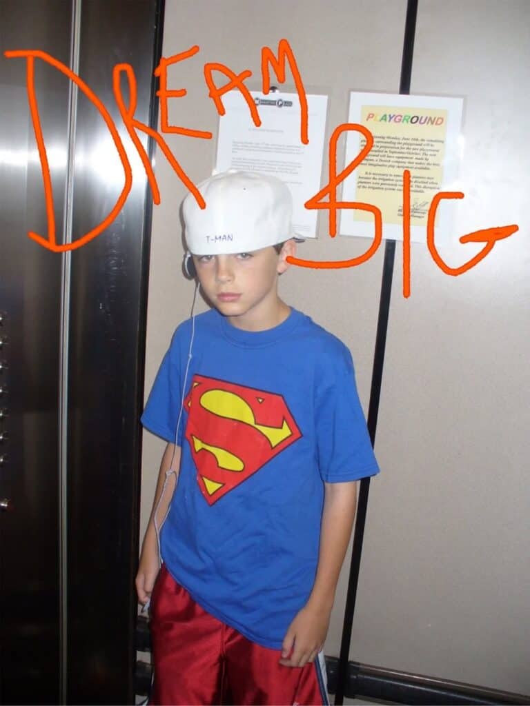 A boy wearing a Superman T-shirt, red shorts, a sideways white cap, and earphones stands in an elevator. Bold orange text over the image reads “DREAM BIG.”.