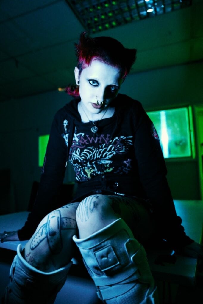 A person with bright red hair, dramatic dark eye makeup, facial piercings, and visible tattoos sits on a table in a dimly lit room with green-tinted windows, wearing a black hoodie and knee braces.