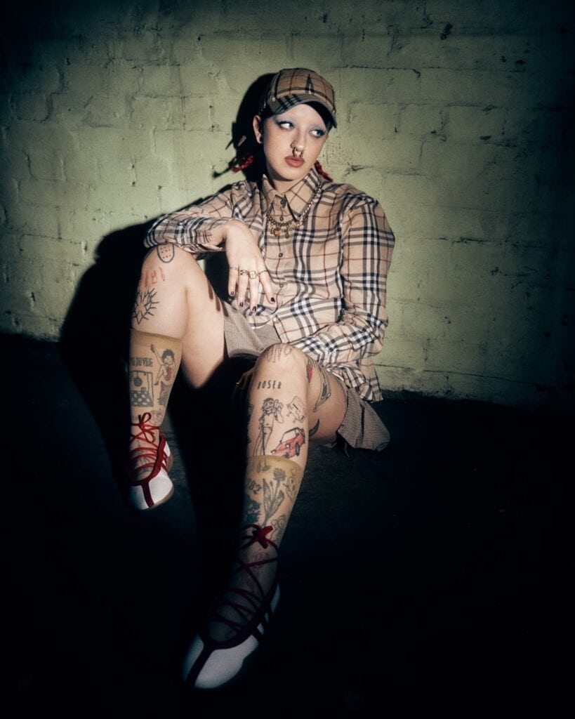 A person wearing a plaid shirt, matching cap, and shorts sits against a light green brick wall. They have visible tattoos on their legs and arms, red lipstick, and are wearing ballet-style shoes with red ribbon laces.
