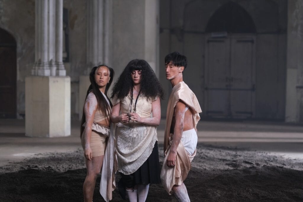 Three people in theatrical costumes stand in a dim, spacious room with columns and arched doors. They face different directions, with neutral expressions, standing on a patch of dark soil for KÁRYYN music video.