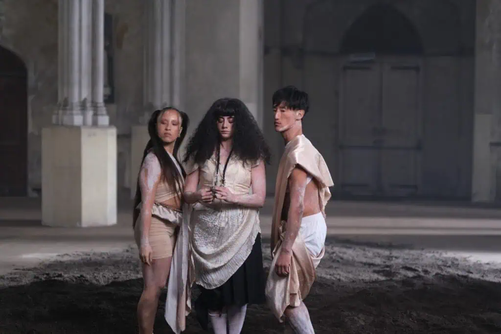 Three people in theatrical costumes stand in a dim, spacious room with columns and arched doors. They face different directions, with neutral expressions, standing on a patch of dark soil for KÁRYYN music video.