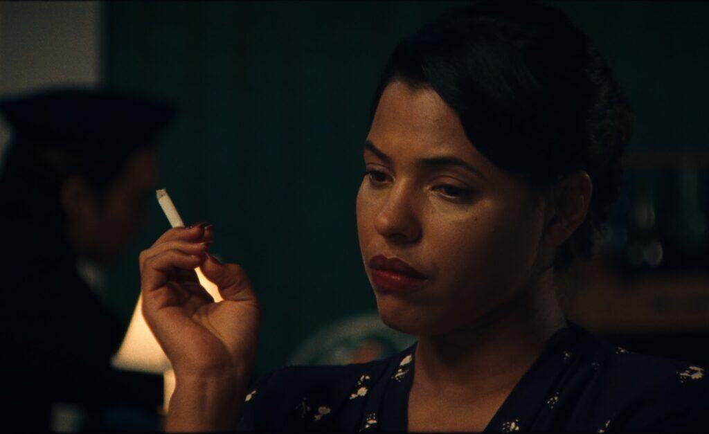 A woman with dark hair and red lipstick holds a cigarette and looks down pensively, evoking the cinematic style of Madeleine Hunt-Ehrlich. The background is dimly lit, with a blurred figure seated behind her.