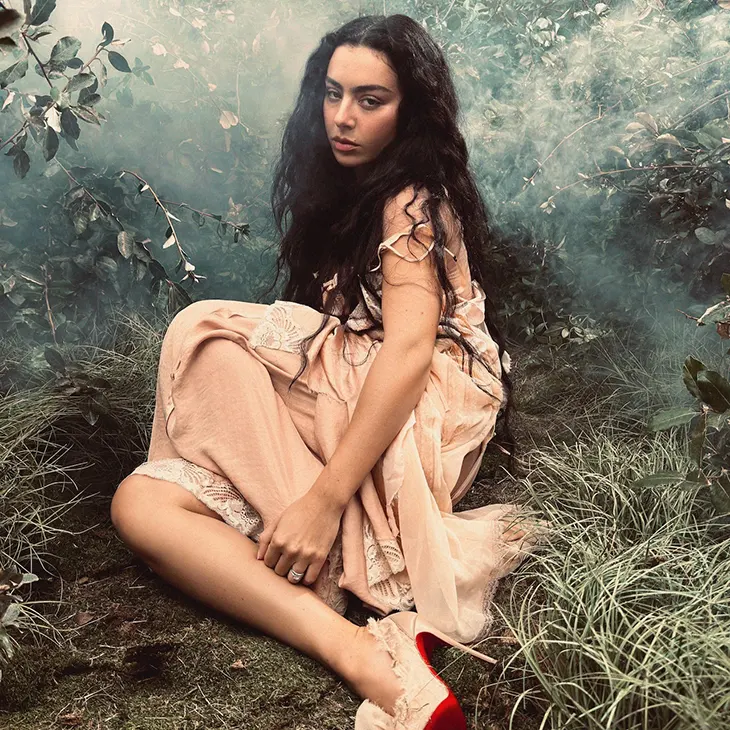 A woman with long dark hair wearing a flowing, pale pink dress sits on the grass in a misty, overgrown forest setting. She looks toward the camera with a calm, distant expression.