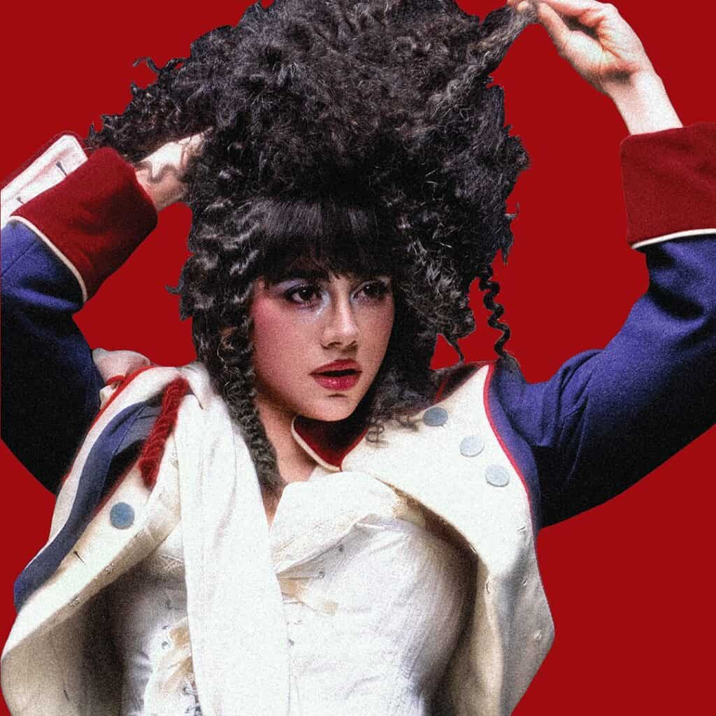 KÁRYYN in a dramatic historical costume with a white jacket and blue sleeves lifts their curly dark hair. They have bold makeup and stand against a solid red background.