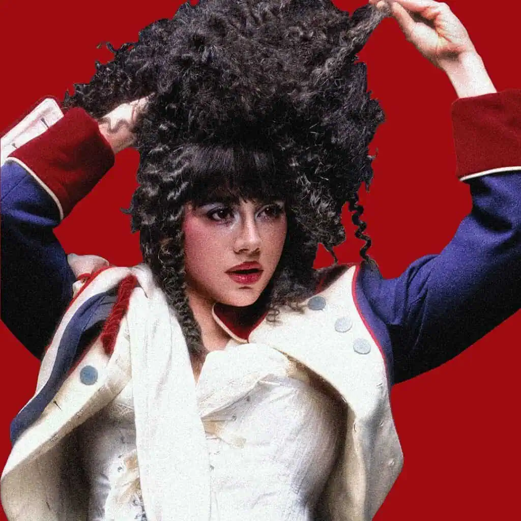 KÁRYYN in a dramatic historical costume with a white jacket and blue sleeves lifts their curly dark hair. They have bold makeup and stand against a solid red background.
