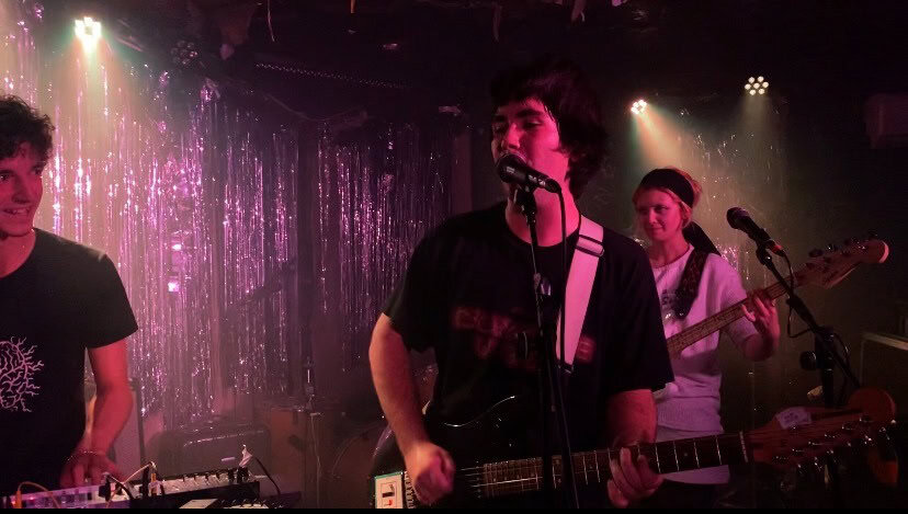 Three musicians perform on stage under pink and purple lights. The guitarist sings into a microphone, another plays bass, and one stands by equipment. Shiny metallic streamers hang as a backdrop, creating a festive atmosphere.