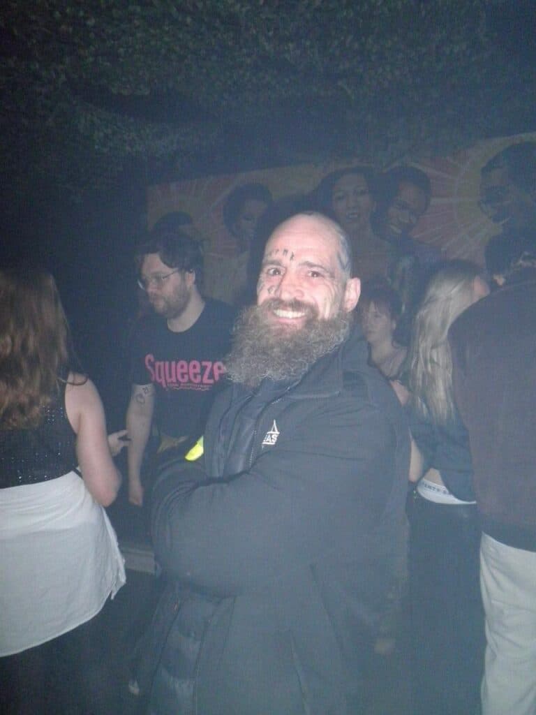 A bearded man with face tattoos smiles at the camera with his arms crossed in a dimly lit, crowded indoor party or club, with people socializing in the background.