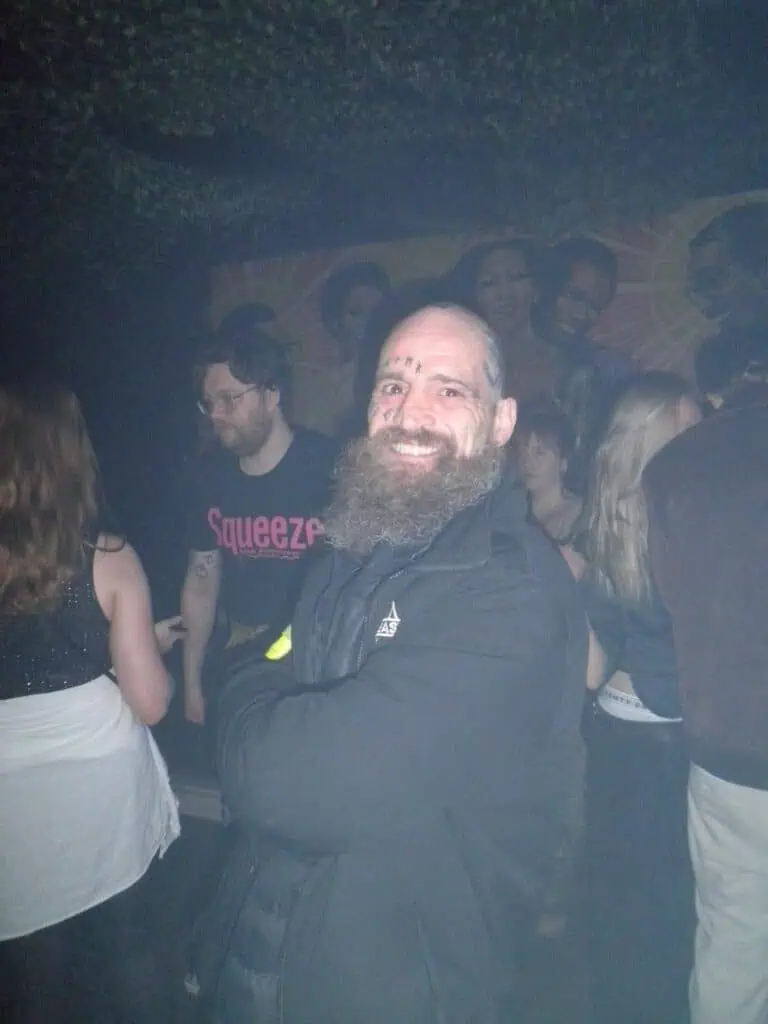 A bearded man with face tattoos smiles at the camera with his arms crossed in a dimly lit, crowded indoor party or club, with people socializing in the background.