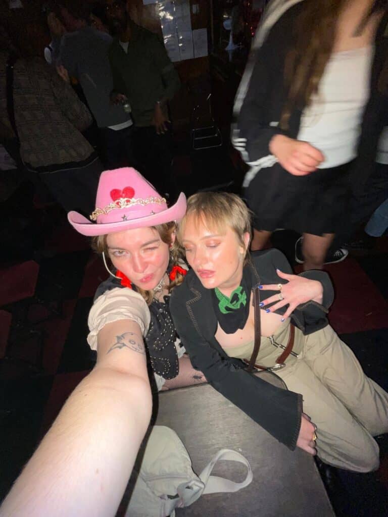 Two people pose for a selfie at a party or club. One wears a pink cowboy hat and winks, while the other wears a green scarf and strikes a pose. There are other people and a dimly lit background.
