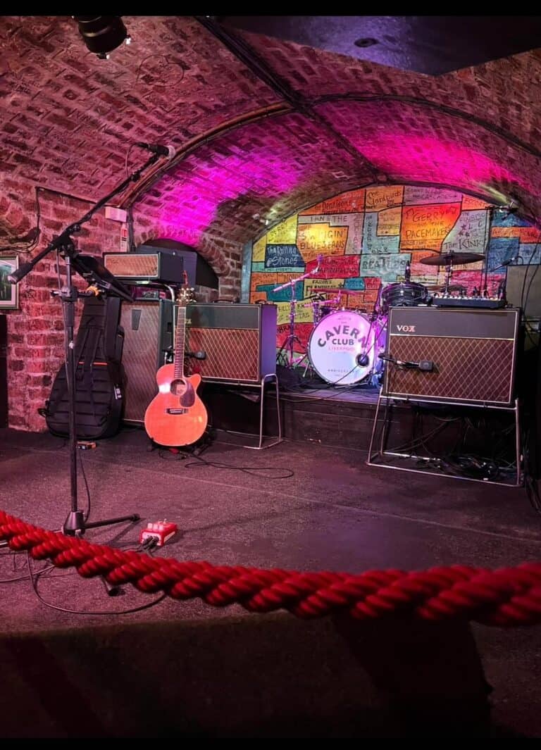 A stage at The Cavern Club with brick arches, colorful wall signs, guitars, VOX amplifiers, a drum set, and a red rope barrier in the foreground, lit by purple and pink lights.